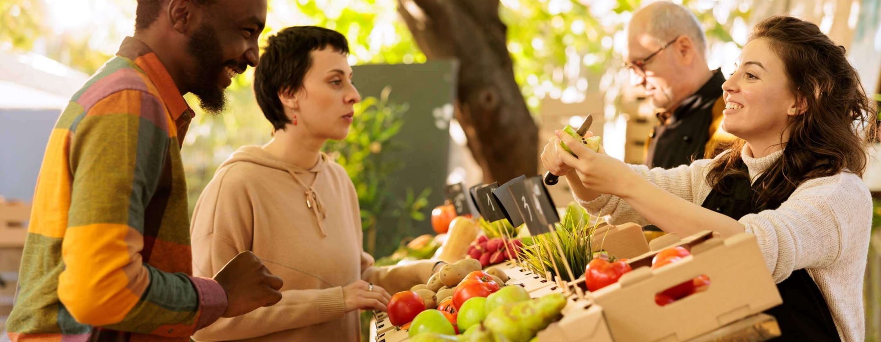 Customers interact with a vendor at an outdoor market stand filled with fresh produce.