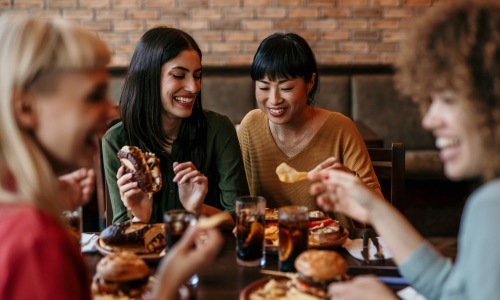 A group of friends enjoying a meal together, smiling and laughing while sharing burgers, fries, and drinks at a restaurant.